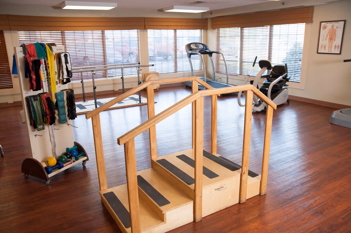 A rehabilitation exercise room with wooden handrails and steps in the center, exercise equipment including a treadmill and stationary bike near large windows with blinds, and a rack holding various weights and resistance bands.