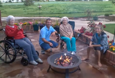 Two elderly women, a younger man, and a young woman sit around a small fire pit on a patio, roasting marshmallows with green fields in the background.
