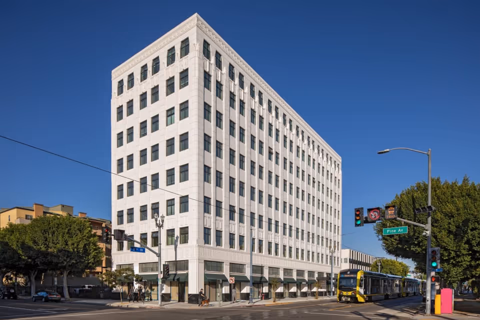 A large white multi-story building on a street corner under a clear blue sky. There are traffic lights, street signs, and a bus on the road. Trees and other buildings are visible in the background.