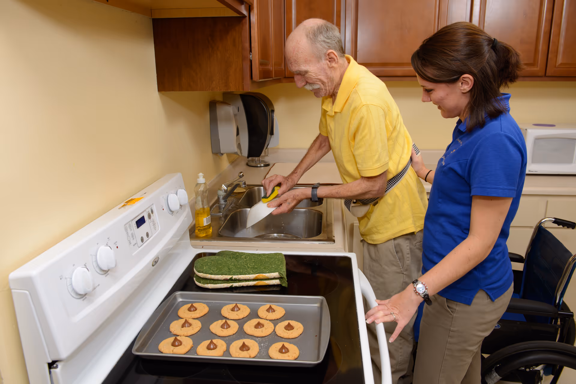 An elderly man washing a baking tray in a kitchen sink while a woman in a blue shirt watches and smiles. There is a tray of freshly baked cookies on the stove, and a wheelchair is visible next to the woman.
