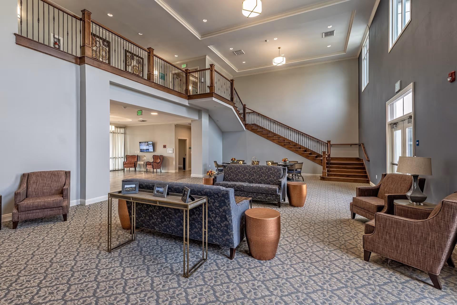 Spacious senior living facility common area with patterned carpet, multiple seating arrangements including armchairs and sofas, a wooden staircase leading to an upper level with railing, large windows letting in natural light, and modern ceiling lights.