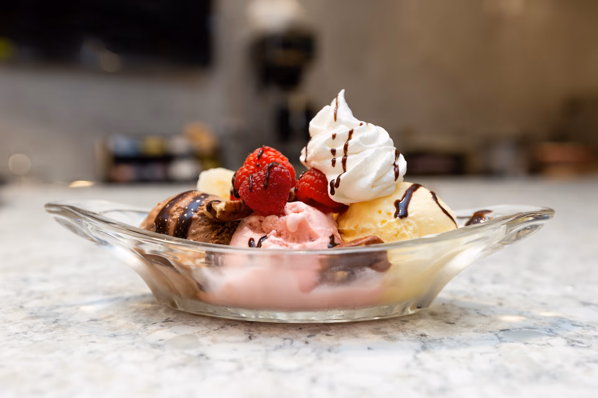 A glass dish containing scoops of chocolate, strawberry, and vanilla ice cream topped with whipped cream, fresh raspberries, and drizzled with chocolate syrup, placed on a marble countertop.