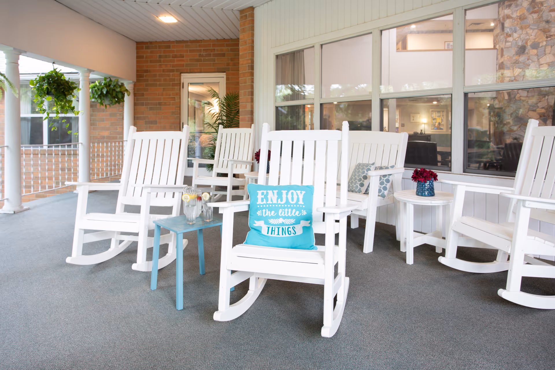 A covered outdoor patio area with white wooden rocking chairs arranged around small tables. One chair has a turquoise pillow with the words 'ENJOY the little THINGS'. There are hanging plants on the left side and large windows on the right side showing an indoor area with furniture.