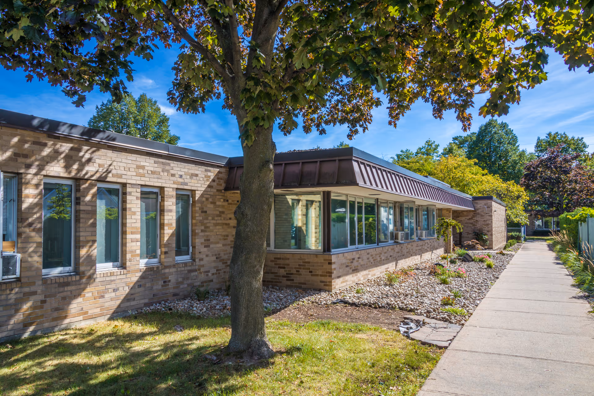 Exterior view of a single-story brick building with multiple windows, a tree in the foreground, and a sidewalk running alongside landscaped areas with rocks and plants under a clear blue sky.