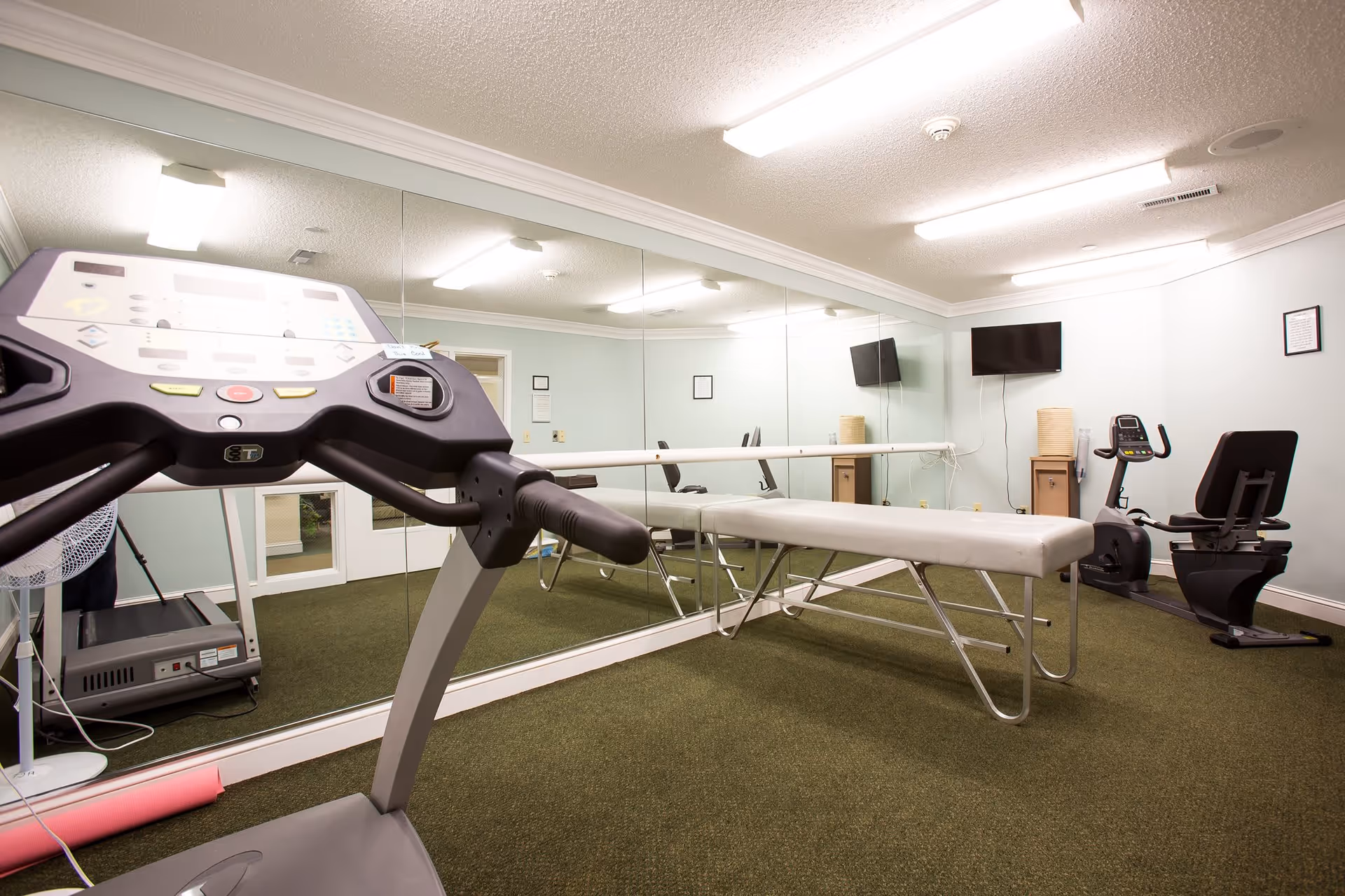 A small fitness room with a treadmill, a recumbent exercise bike, a padded exercise table, and a large wall mirror. The room has light-colored walls, a green carpet, and two wall-mounted televisions.