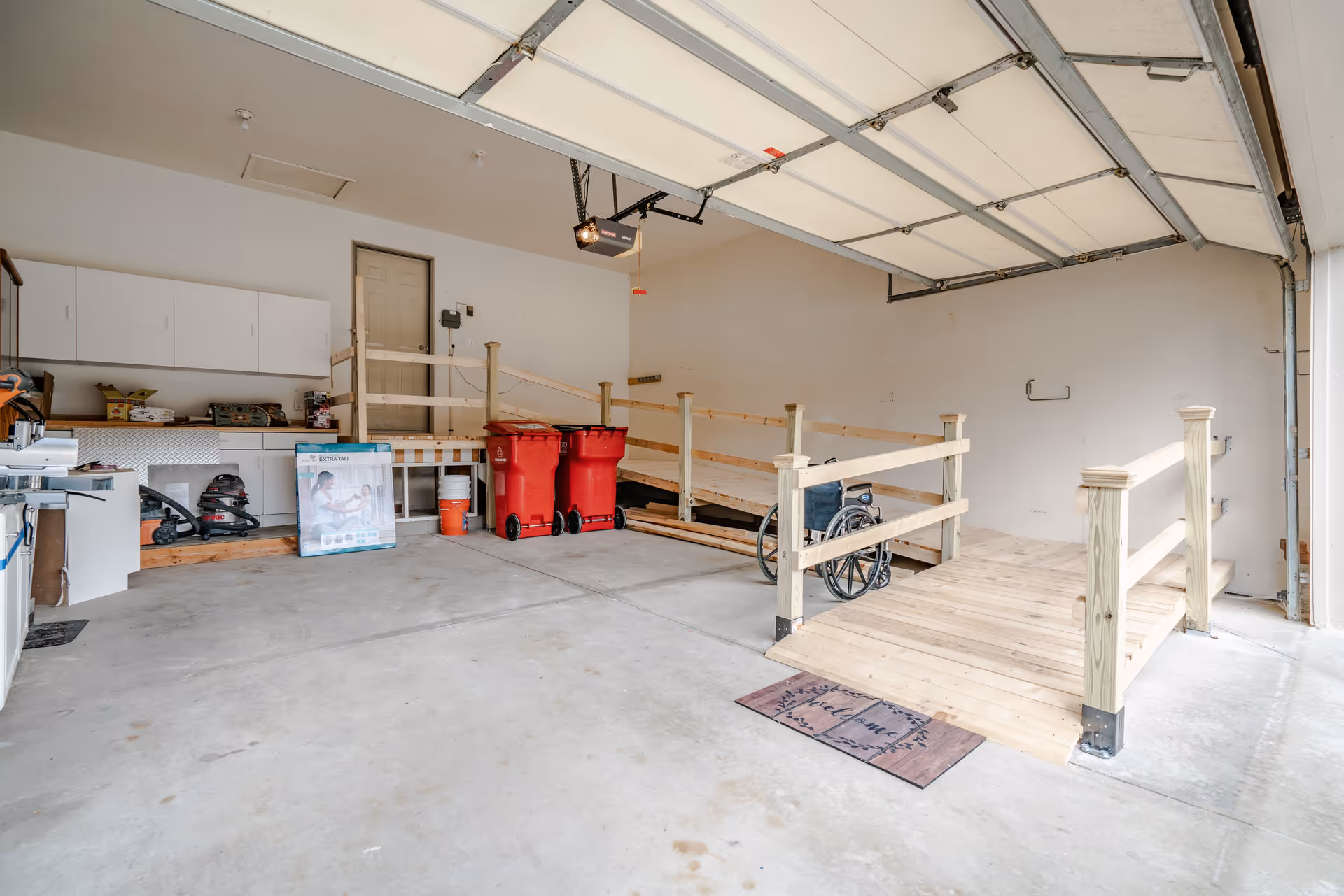 Interior view of a garage with a concrete floor, a wooden ramp with railings leading to a door, a wheelchair positioned on the ramp, two red trash bins, white cabinets mounted on the wall, and various cleaning and maintenance supplies.