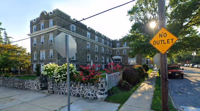 Exterior view of a multi-story stone building with a garden featuring white and pink flowers in front, a stone fence, a sidewalk, and a street with a 'No Outlet' sign and parked cars.