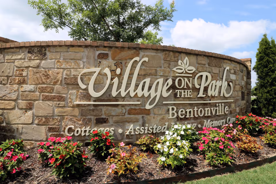 Stone wall sign for Village on the Park Bentonville with flowers planted in front and a tree and blue sky in the background.