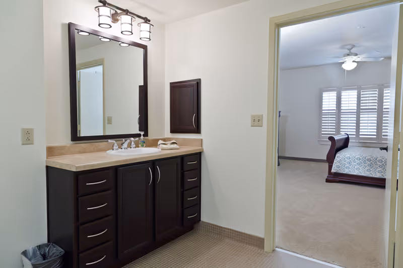 Bathroom vanity with a large mirror, sink, and dark wood cabinets. A doorway leads to a bedroom with a bed, ceiling fan, and window with white shutters.