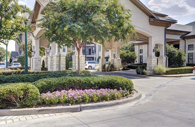 Exterior view of Conservatory At Keller Town Center showing a covered entrance with stone pillars, landscaped bushes, a tree, and a flower bed with purple flowers. Several cars are parked nearby under a partly cloudy sky.