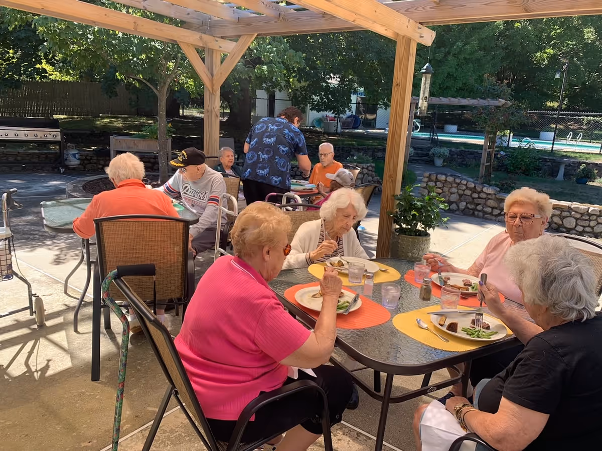 A group of elderly people sitting at outdoor tables under a wooden pergola, eating a meal together. The setting is a sunny patio area with trees and a stone wall in the background. A caregiver is assisting one of the residents at a table further back.