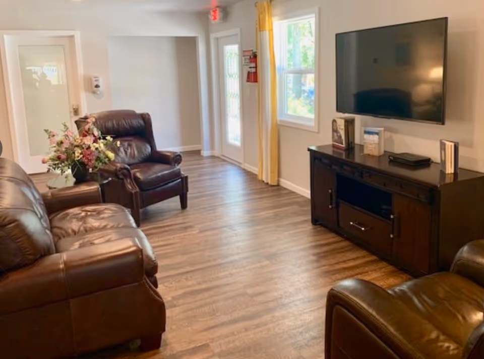 A cozy living room with brown leather armchairs and a sofa arranged around a dark wooden TV stand with a flat-screen television mounted on the wall. There is a window with yellow curtains and a door with a glass panel. A vase with flowers is placed on a small side table next to one of the armchairs. The room has wooden flooring and light-colored walls.