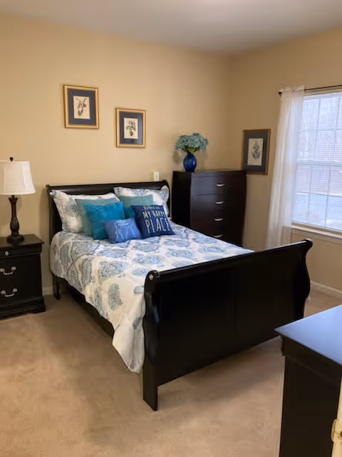A neatly made bed with a black wooden frame in a bedroom. The bed has white and blue patterned bedding with several decorative pillows, including one that says 'My Happy Place'. There are two framed pictures on the beige wall above the bed. To the left of the bed is a black nightstand with a white lamp. To the right is a black dresser with a blue vase holding flowers. A window with white curtains lets in natural light.