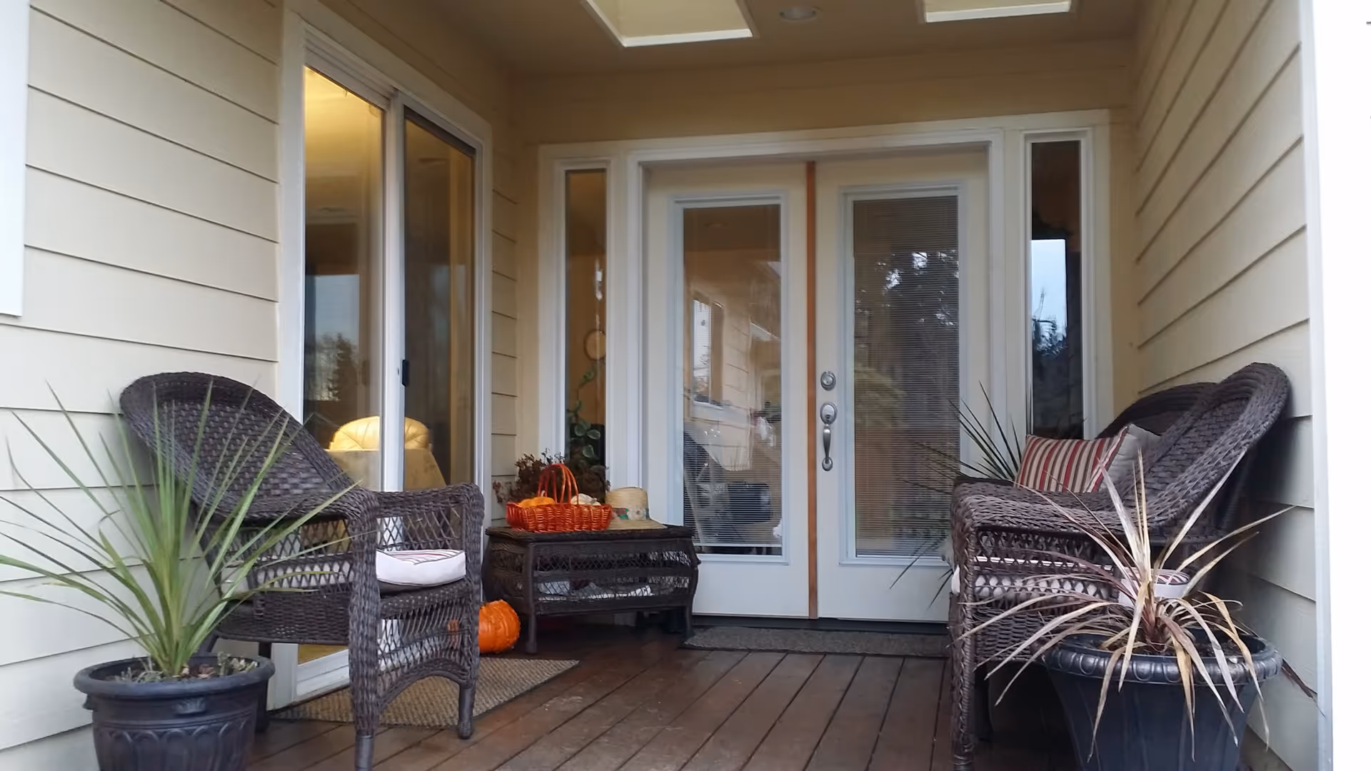 A covered outdoor porch area with wooden flooring, featuring two wicker chairs with cushions, a wicker loveseat with a striped pillow, two potted plants, a small wicker table with a basket and decorative items, and glass double doors leading inside.