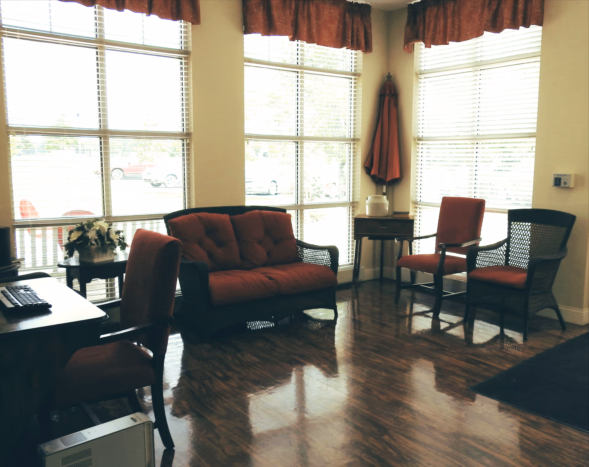 A bright sitting area with large windows covered by blinds and red valances. The room features a red cushioned loveseat, two red cushioned chairs, a small wooden table with a white decorative item, and a desk with a computer keyboard and chair. The floor is polished wood, and there is an umbrella in the corner near the windows.