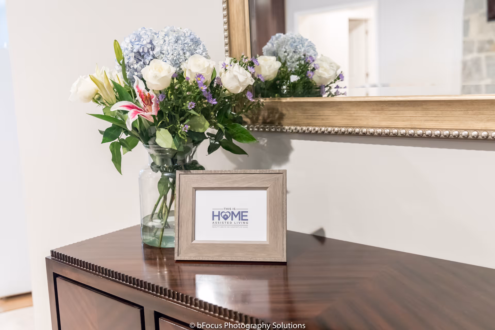 A wooden table with a glass vase holding a bouquet of flowers including white roses, lilies, and purple flowers. A framed sign on the table reads 'This is Home Assisted Living'. A large decorative mirror hangs on the wall behind the table.