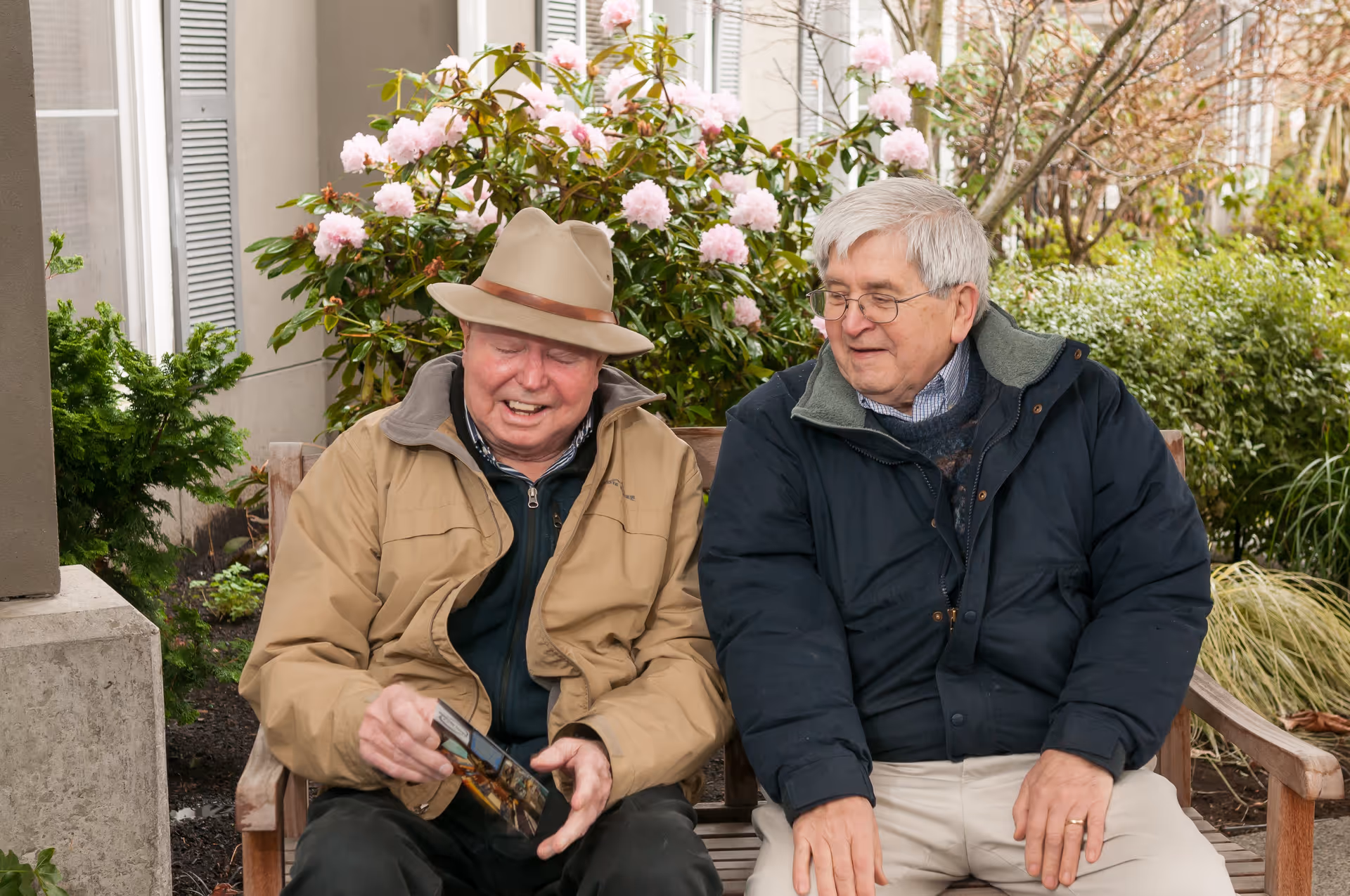 Two elderly men sitting on a wooden bench outdoors, engaged in conversation. One man is wearing a tan jacket and a beige hat, holding a postcard or photo, while the other man is wearing glasses and a dark jacket. Behind them are green bushes and blooming pink flowers.