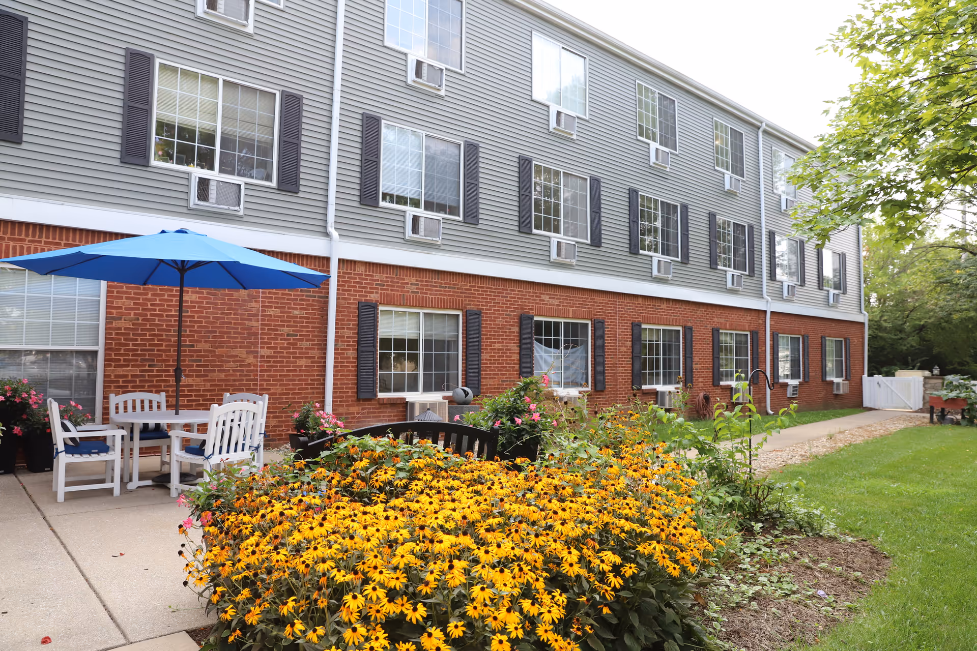 Outdoor garden area at Rosewalk at Lutherwoods with a large bed of yellow flowers, a black bench, a patio table with white chairs and a blue umbrella, and a three-story building with brick and gray siding in the background.