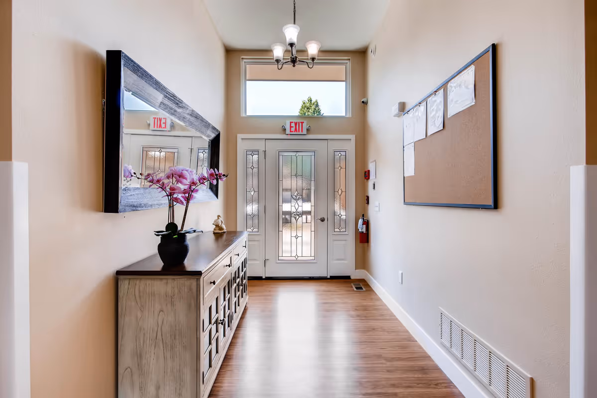 Bright entry hallway with a console table and mirror on the left, a corkboard on the right, and a decorative glass front door under an EXIT sign.