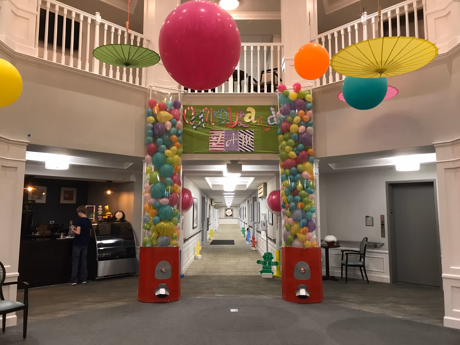 Interior view of a senior living facility lobby decorated with colorful balloons inside two tall transparent columns resembling gumball machines. Large colorful hanging paper umbrellas and balloons are suspended from the ceiling. A green banner with colorful letters is displayed above the columns. A person is standing near a counter on the left side, and there is an elevator on the right side.