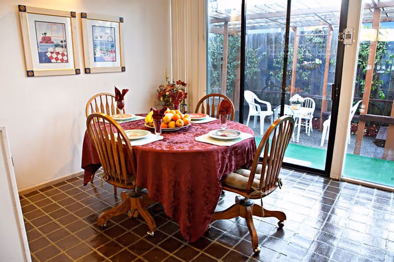 A dining room with a round wooden table covered with a red tablecloth set for four people with plates, glasses, and napkins. A centerpiece bowl of fruit is on the table. Four wooden chairs with cushions surround the table. Two framed pictures hang on the wall, and a sliding glass door opens to an outdoor patio area with white plastic chairs and a small table.