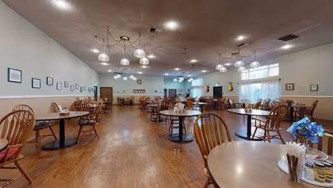 A spacious dining room with multiple round wooden tables and chairs arranged neatly. The room has wooden flooring, light-colored walls adorned with framed pictures, and several hanging light fixtures. There are windows on the far wall allowing natural light to enter, and a few decorative items are visible on the walls and shelves.