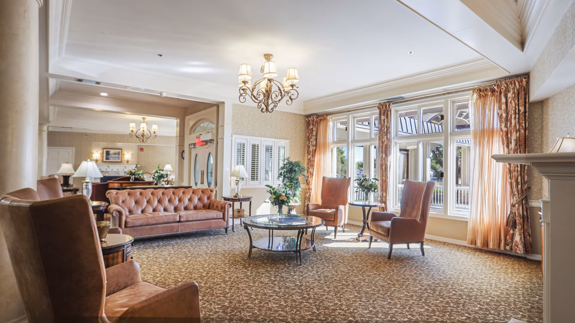 A bright and spacious living room area in a senior living facility with large windows letting in natural light. The room features brown leather sofas and armchairs arranged around a glass coffee table with a flower arrangement. There are elegant chandeliers hanging from the ceiling, patterned carpet flooring, and floral curtains on the windows. The room has a warm and inviting atmosphere with lamps and plants adding to the decor.