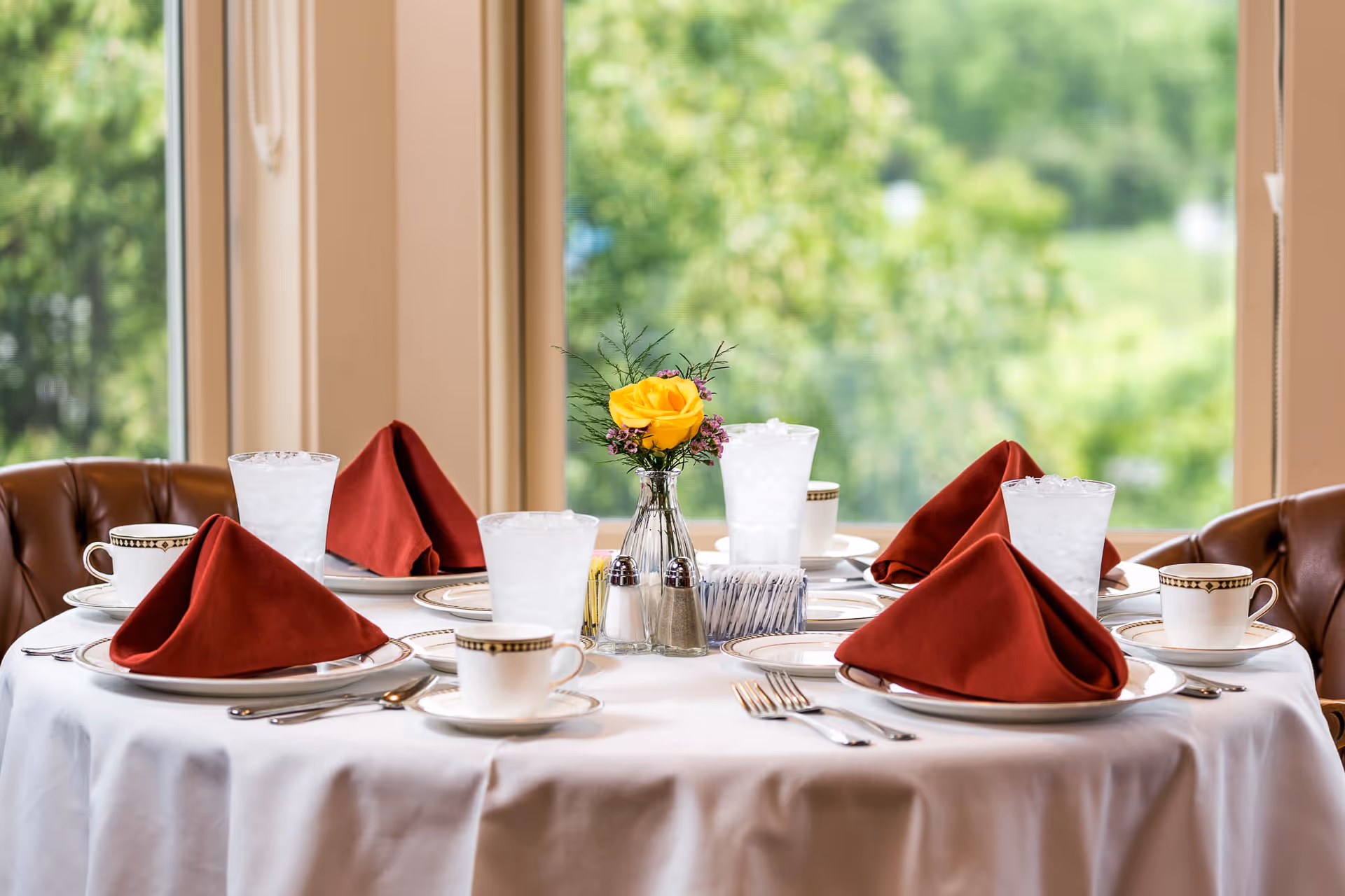 Round dining table set for four with red folded napkins, china, iced water glasses and a yellow rose centerpiece in front of large windows.