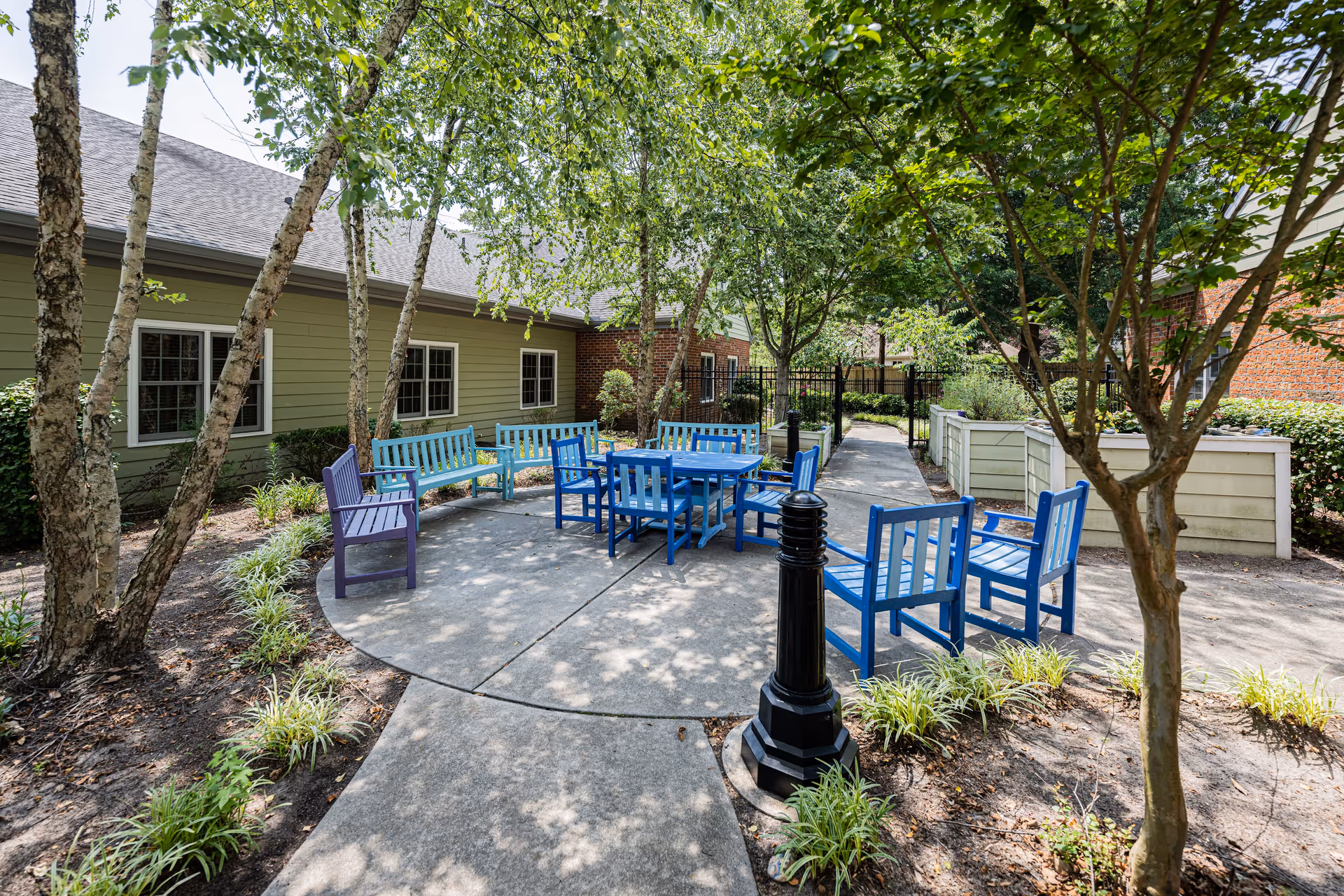 Outdoor seating area in a garden courtyard with blue and purple benches and chairs arranged around a blue table. The area is surrounded by trees, plants, and a paved walkway, with a building visible in the background.