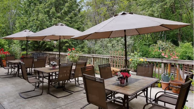 Outdoor patio area with multiple tables and chairs under large beige umbrellas. The patio is surrounded by wooden railing and lush green trees and plants, with potted flowers and gardening tools visible along the railing.