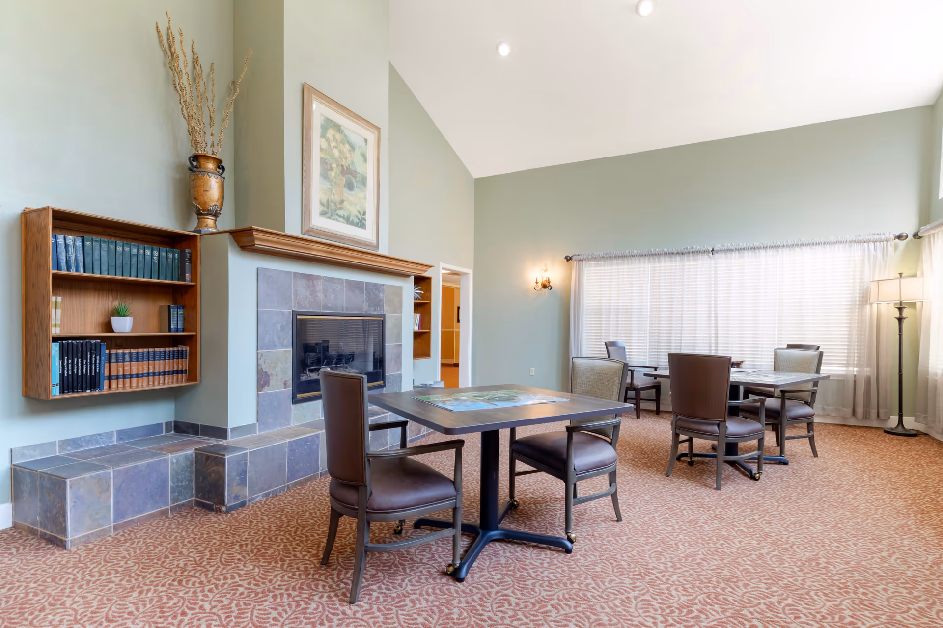A spacious room with a high ceiling painted in light green, featuring a fireplace with a tiled surround and a wooden mantel. There is a wooden bookshelf mounted on the wall with books and a small plant. The room has several tables with chairs arranged around them, a large window with sheer curtains, a floor lamp, and a wall sconce providing light.