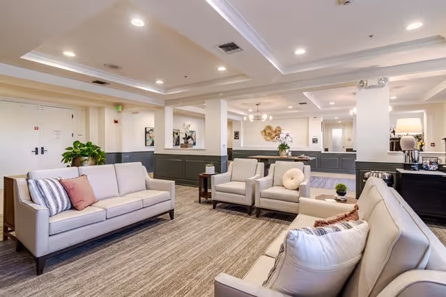 A spacious and well-lit living room area in a senior living facility featuring two beige sofas with decorative pillows, two beige armchairs, a side table with a plant, and a carpeted floor. The room has white walls with dark green wainscoting, recessed ceiling lights, and decorative wall art. There is a coffee station with a lamp and coffee maker on the right side.