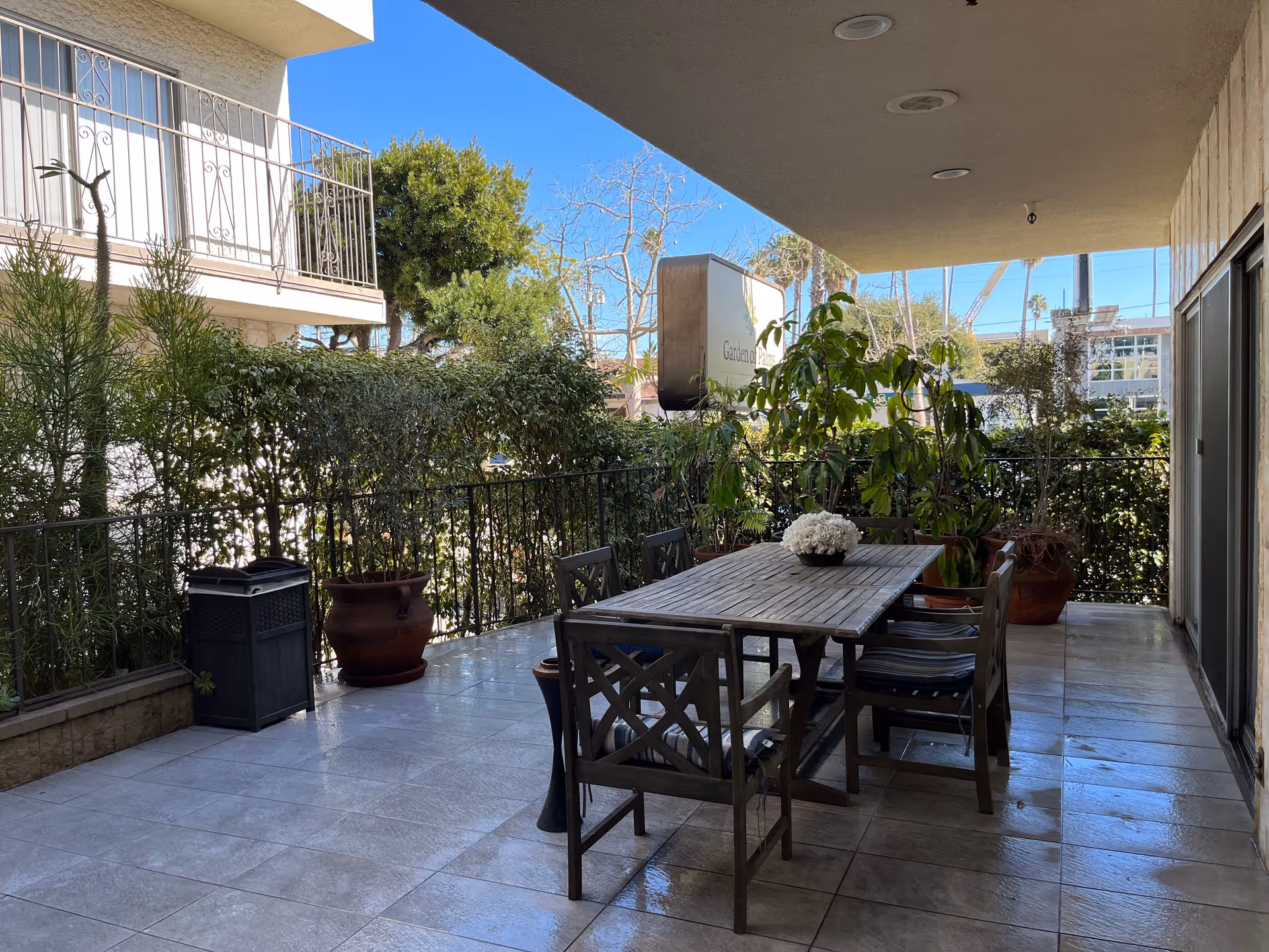 Outdoor covered patio area with a wooden dining table and six chairs. The table has a small flower arrangement in the center. Surrounding the patio are potted plants and greenery, with a clear blue sky visible in the background.