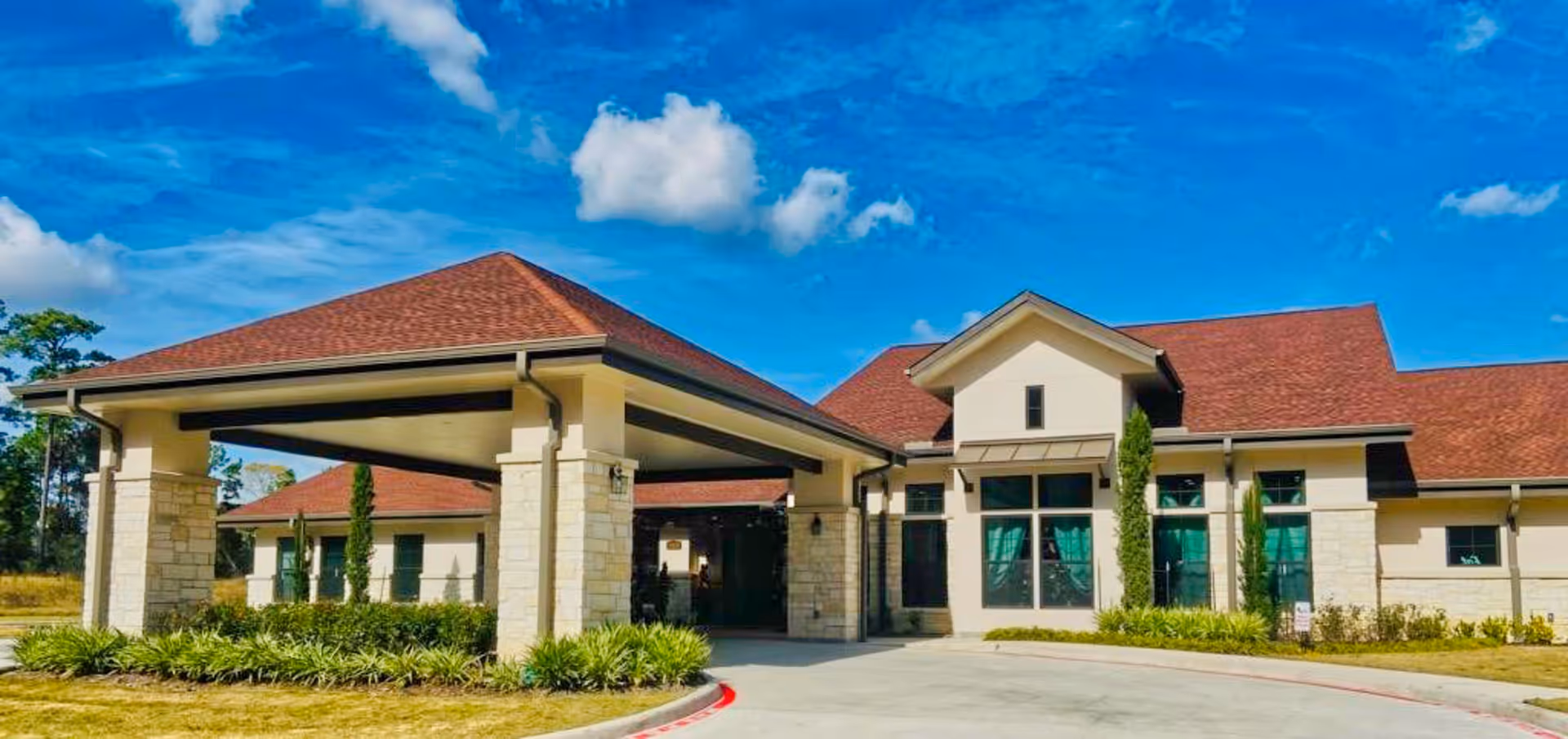Front entrance of a single-story red-roofed assisted living building with a covered porte-cochere, windows, and landscaping under a blue sky.
