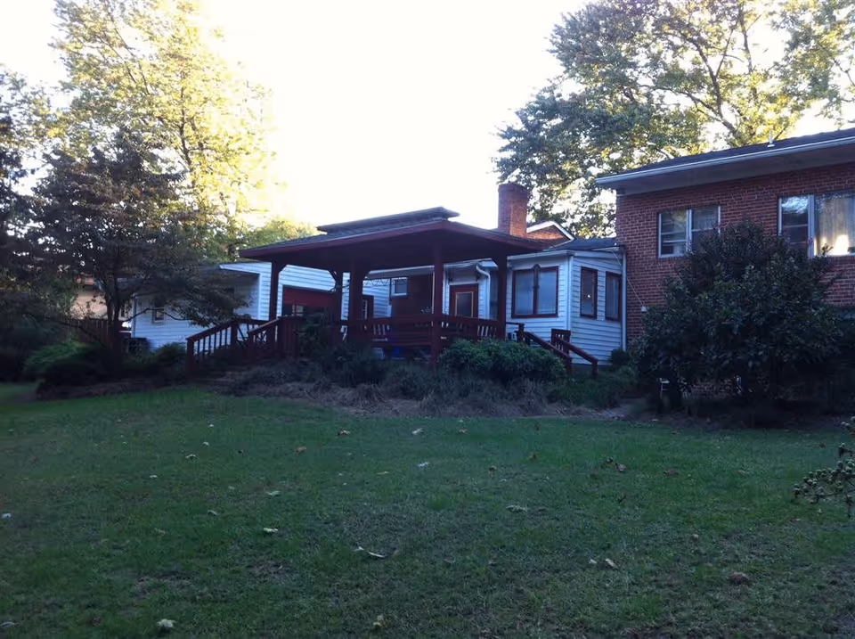 Exterior view of a senior living facility showing a grassy yard with trees and bushes. The building has a combination of brick and white siding with a covered wooden porch and steps leading up to it.