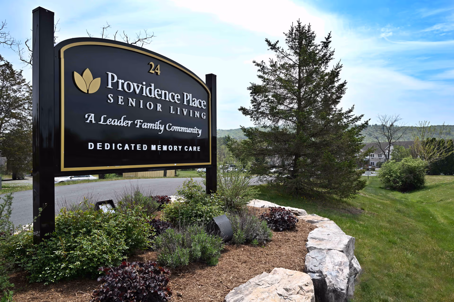 Outdoor view of a large black and gold sign for Providence Place Senior Living, a Leader Family Community with dedicated memory care, surrounded by landscaped greenery and rocks with trees and a road in the background under a partly cloudy sky.