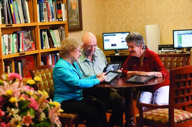 Three elderly individuals sitting around a round wooden table in a cozy room with bookshelves and computers in the background. They are engaged with tablet devices, smiling and interacting with each other. The room has warm lighting and patterned wallpaper.
