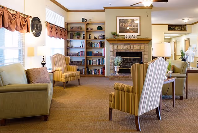 A cozy living room area with a green sofa, two striped armchairs, a wooden bookshelf filled with books and decorative items, and a stone fireplace with a framed picture and the word 'HOME' on the mantel. The room has beige carpet, cream-colored walls with wooden trim, and windows with brown valances letting in natural light.