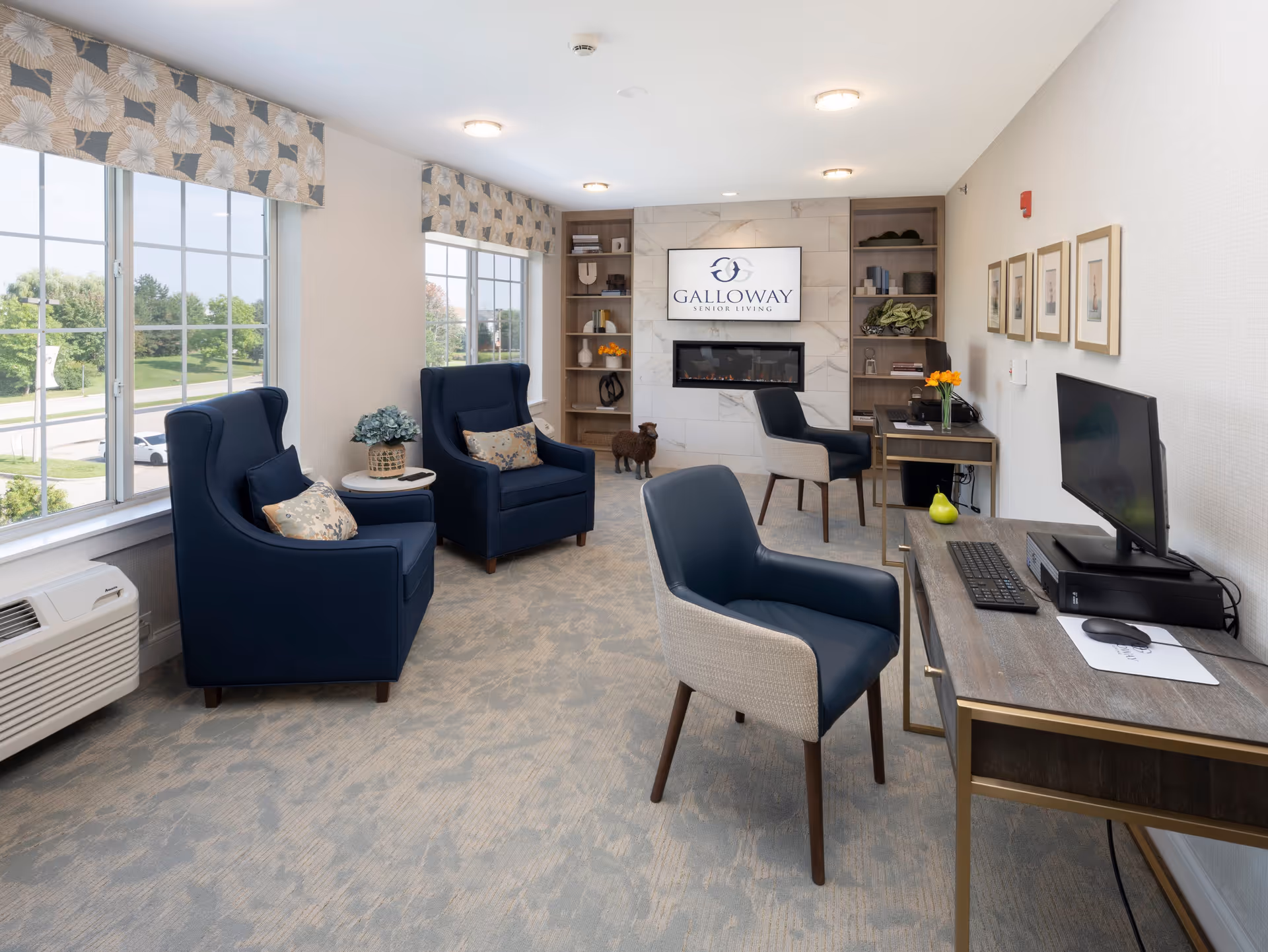 A bright and modern senior living common area with large windows, two navy blue armchairs with patterned pillows, a small round table with a plant, two desks with chairs and computer monitors, a decorative fireplace with a TV above displaying the Galloway Senior Living logo, built-in shelves with decorative items, and framed pictures on the wall.