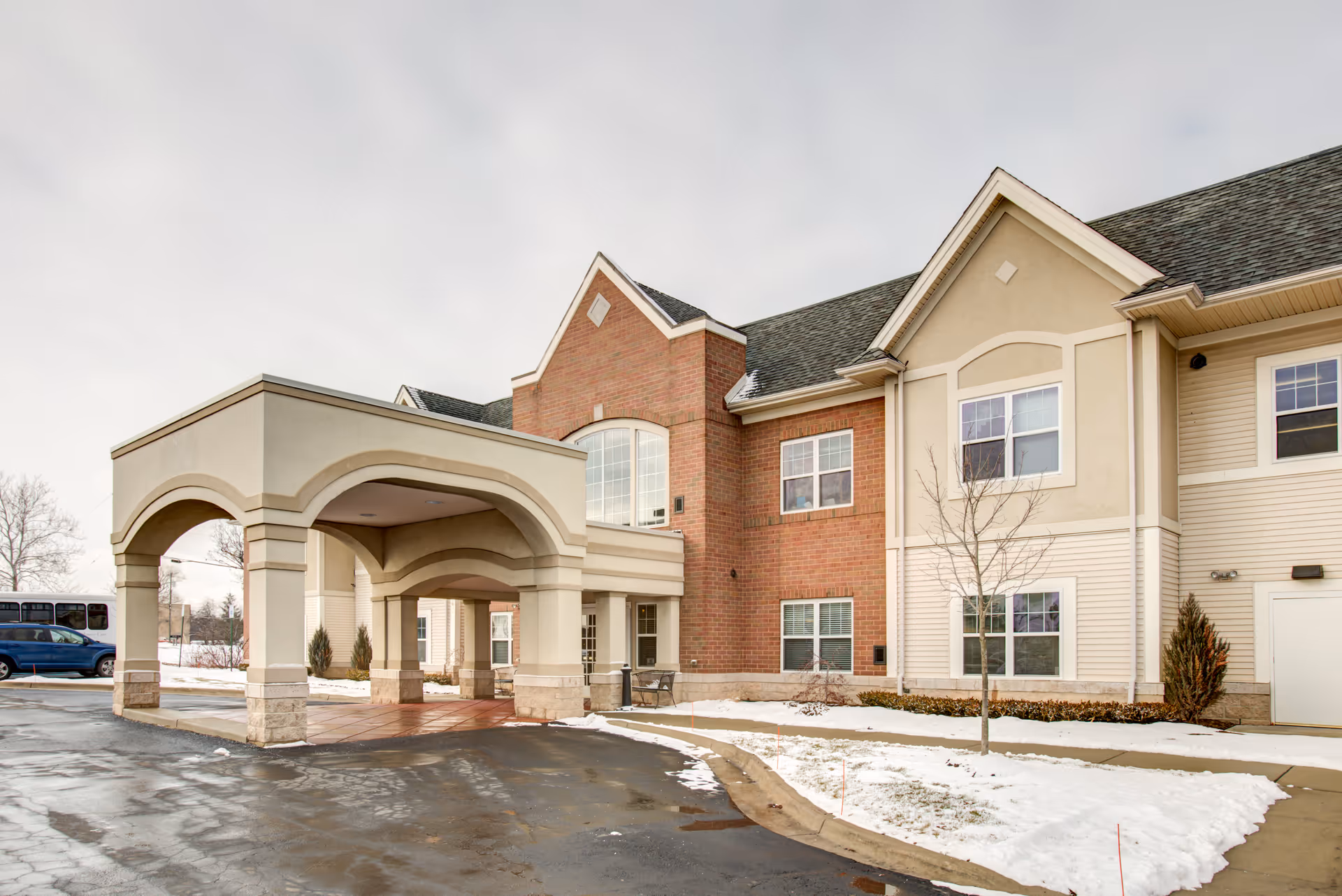 Exterior view of a senior living facility building with a covered entrance, brick and beige siding, snow on the ground, and a cloudy sky.