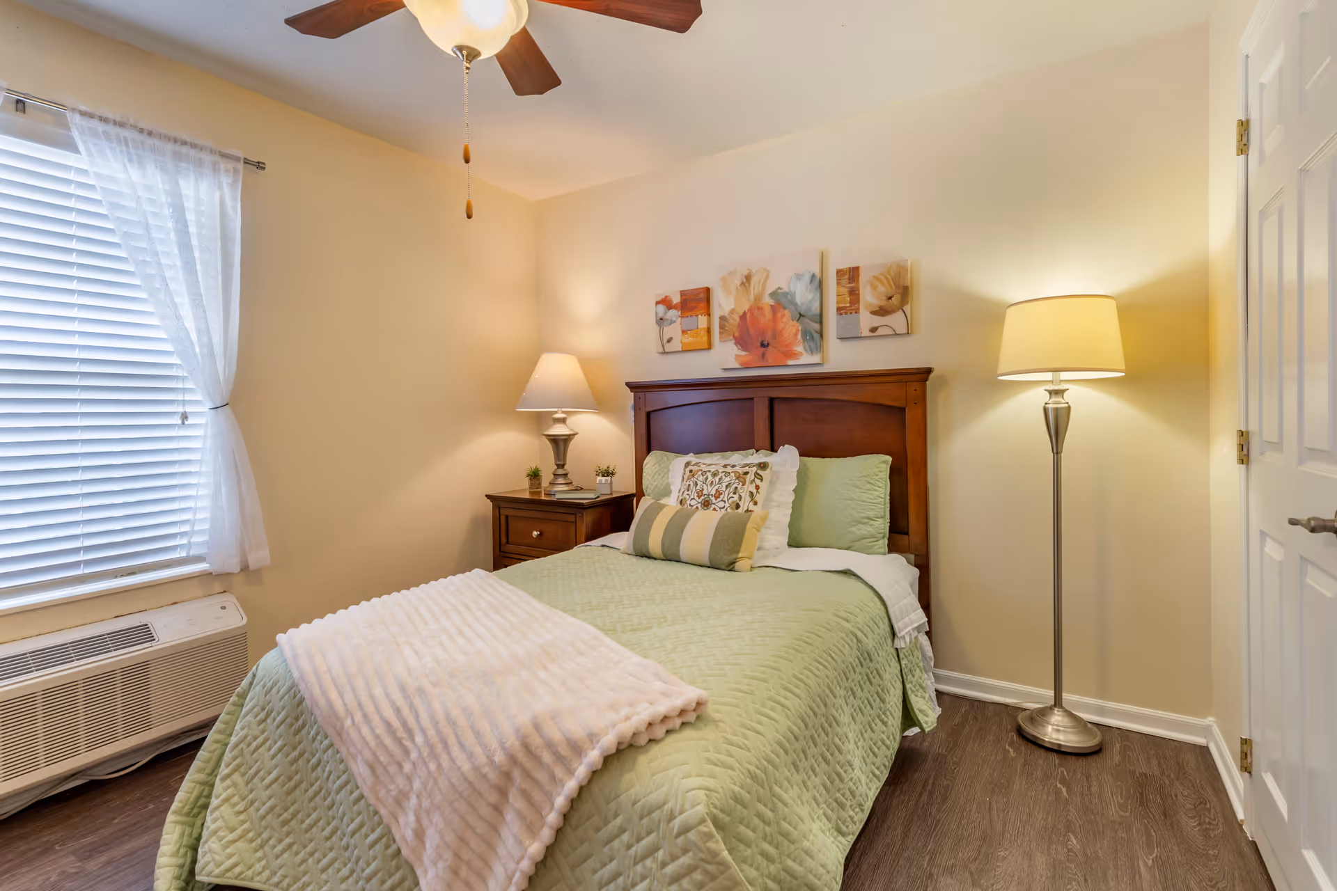 Cozy furnished bedroom with a made bed, wooden headboard, nightstand and lamps, a window with blinds, and a ceiling fan.