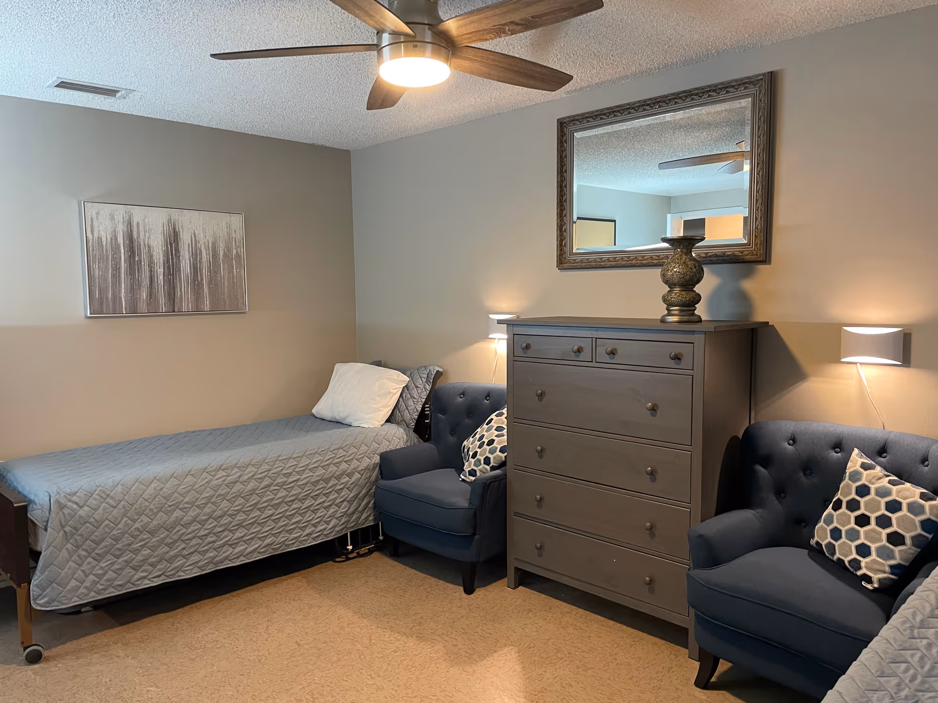 A neat bedroom with a single bed, two blue tufted armchairs, a gray chest of drawers topped by a mirror and a ceiling fan.