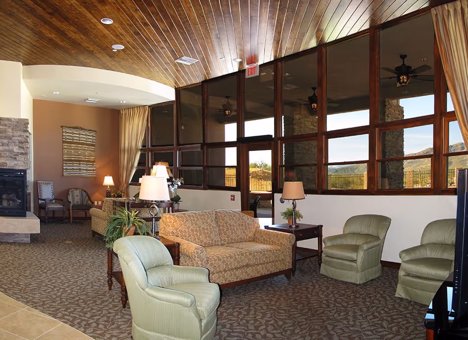 A cozy senior living facility lounge area with patterned carpet, a floral-patterned sofa, two green armchairs, wooden side tables with lamps, a stone fireplace, and large windows showing an outdoor view of mountains and a patio.