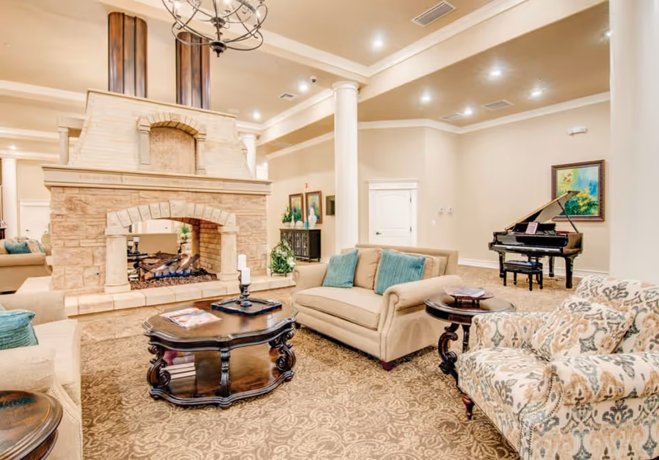 A spacious and elegant living room area featuring a large stone fireplace with a fire burning, beige sofas with teal cushions, a patterned armchair, a dark wooden coffee table, and a grand piano in the background. The room has beige walls, carpeted floors, white columns, and recessed lighting in the ceiling.