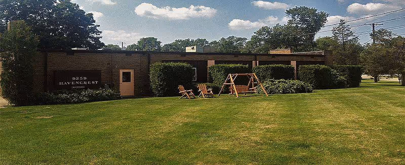 Single-story brick building with a sign reading '9259 HAVENCREST' on the front wall, surrounded by green bushes and a large grassy lawn. There are wooden outdoor chairs and a swing set on the lawn, with trees and a partly cloudy sky in the background.