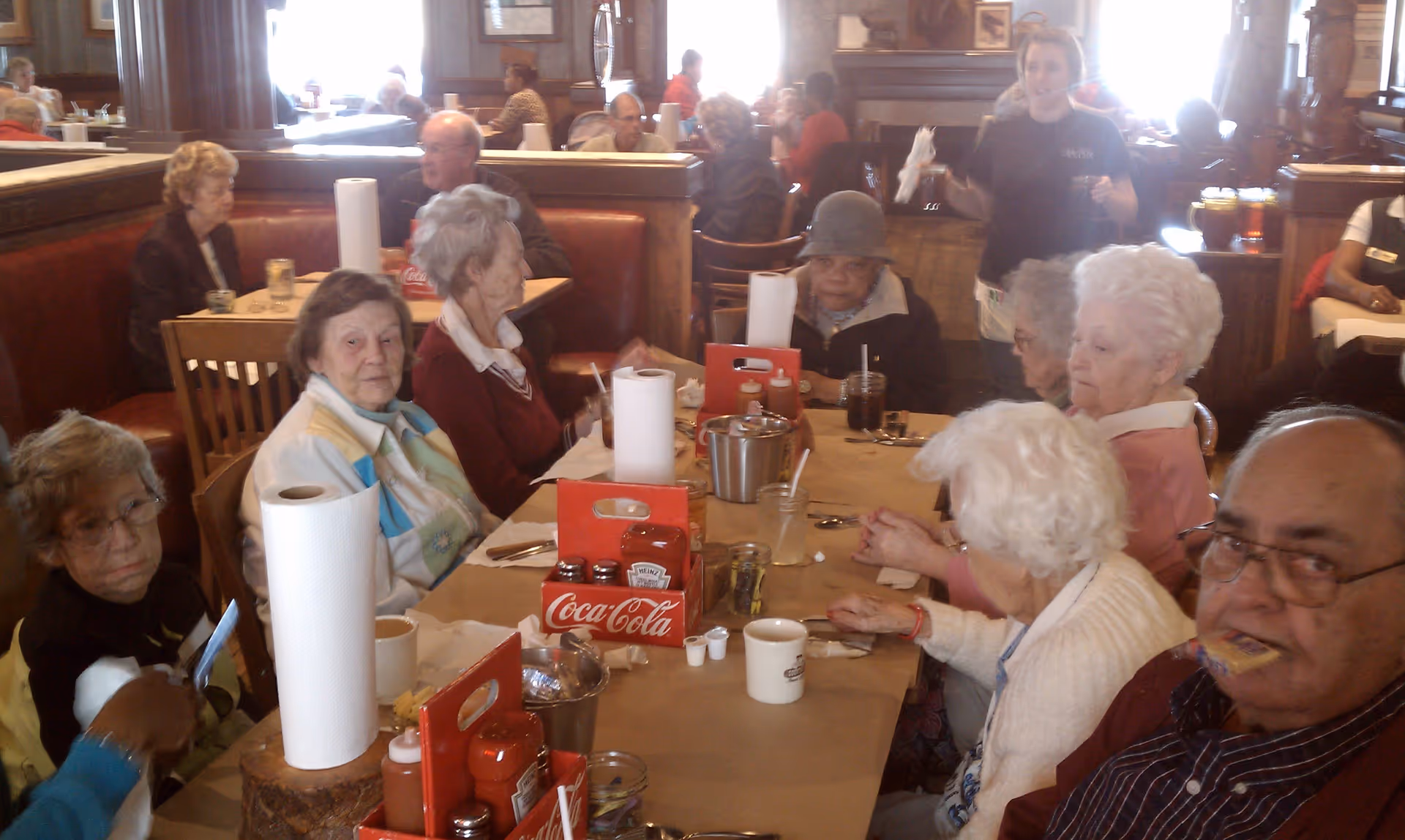 A group of elderly people sitting around a long table in a restaurant or dining area, with condiments, paper towels, cups, and utensils on the table. A waitress is standing in the background holding napkins and a drink. The setting appears warm and busy with other diners in the background.