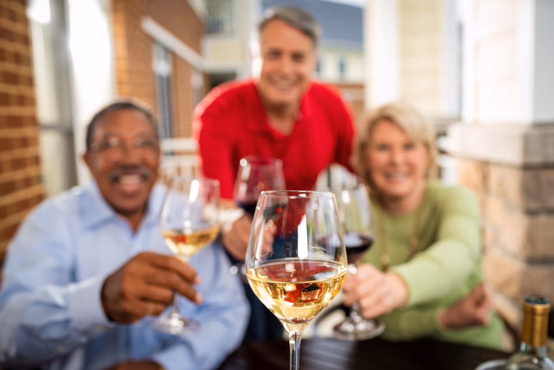 Three smiling adults sitting outdoors on a patio, holding and clinking wine glasses in a celebratory toast.