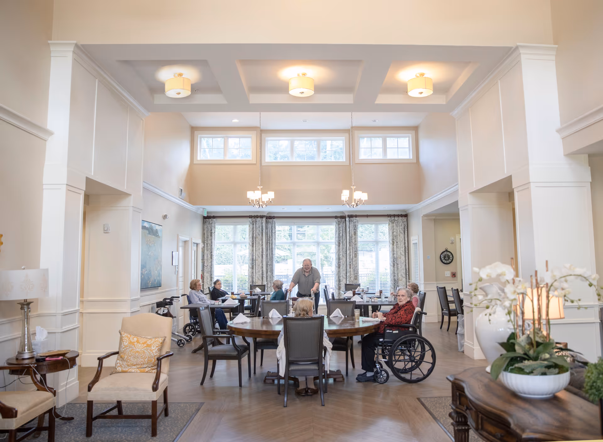 A bright and spacious dining room in a senior living community with several elderly residents seated around a large round table. The room features high ceilings with recessed lighting, large windows with curtains allowing natural light, and elegant decor including a lamp, artwork, and a floral arrangement on a side table.