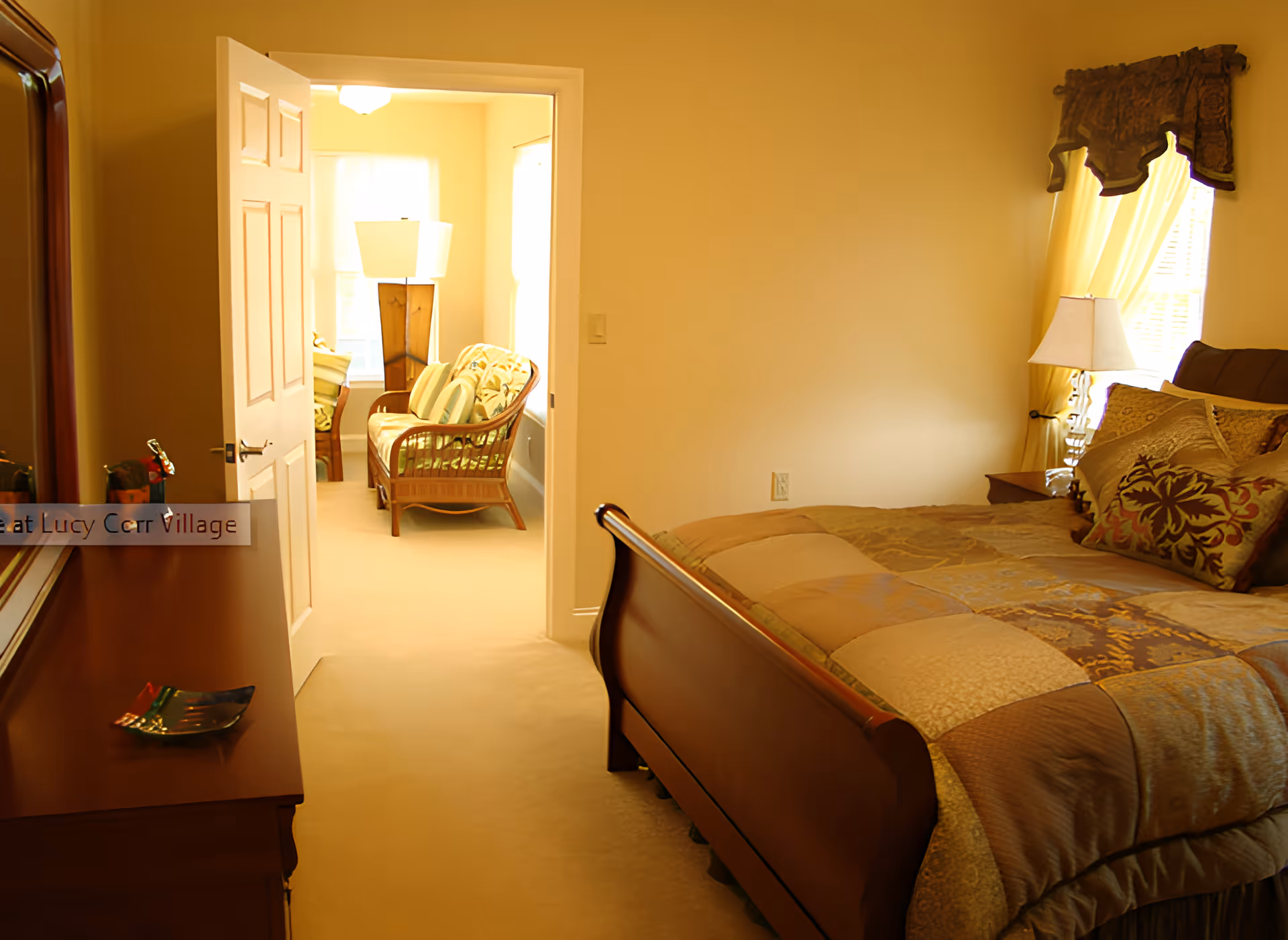 A warmly lit bedroom with a wooden bed covered in a patchwork quilt and decorative pillows. To the right, there is a window with curtains and a bedside table with a lamp. An open door leads to a sunlit sitting area with a wicker loveseat and a floor lamp.