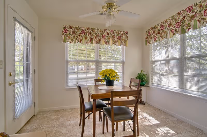 A bright dining area with a wooden table and four chairs. A pot of yellow flowers is centered on the table. The room has large windows with floral valances and blinds, allowing natural light to fill the space. A ceiling fan with a light fixture is mounted above the table. There is a door with blinds on the left side and a small side table with a green plant in the corner.